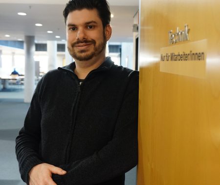 Eine Person an eine Tür gelehnt. Auf der Tür ist die Aufschrift "Technik. Nur für Mitarbeiter/innen" zu lesen. Hinter der Person kann man in einen Lesesaal blicken.