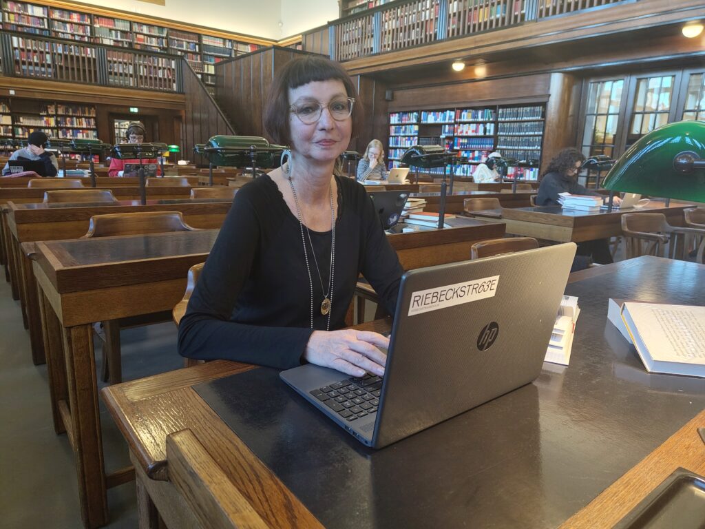 Eine Frau sitzt an einem Schreibtisch aus Holz. Vor ihr steht ein aufgeklapptes Notebook, um das mehrere aufgeschlagene Bücher verteilt sind. Im Hintergrund sieht man einen großen Raum, in dem weitere Schreibtische hintereinander aufgereiht stehen. Hier sitzen weitere Leute und lesen oder arbeiten. Die rückwärtige Wand besteht aus einem großen Bücherregal mit vielen alt aussehende Medienwerken. In der Rechten Bildhälfte befindet sich eine verglaste Eingangstür. Auch hier ist die Architektur älter.  