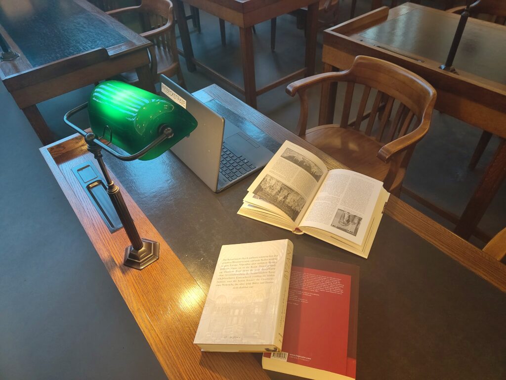 Man sieht von oben auf einen alten Schreibtisch aus Holz.  Davor steht ein dunkler Stuhl mit geschwungener Lehner, der ebenfalls aus Holz ist. Auf dem Tisch stehen ein aufgeklapptes Notebook, eine grüne, altertümliche Leselampe und mehrere aufgeschlagene Bücher.