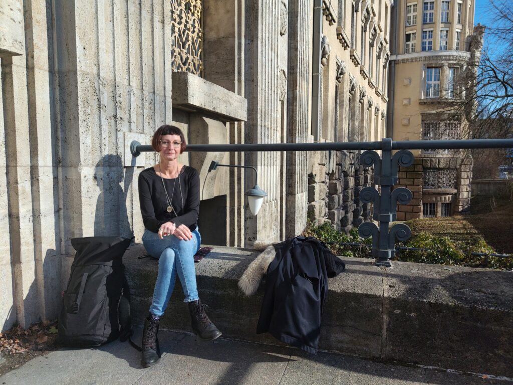 Eine Frau sitzt vor der sandsteinfarbenen Fassade eines historischen Gebäudes. Die Sonne scheint. Im Hintergrund sieht man ein rundes Erker des Gebäudes mit vielen Sprossenfenstern,