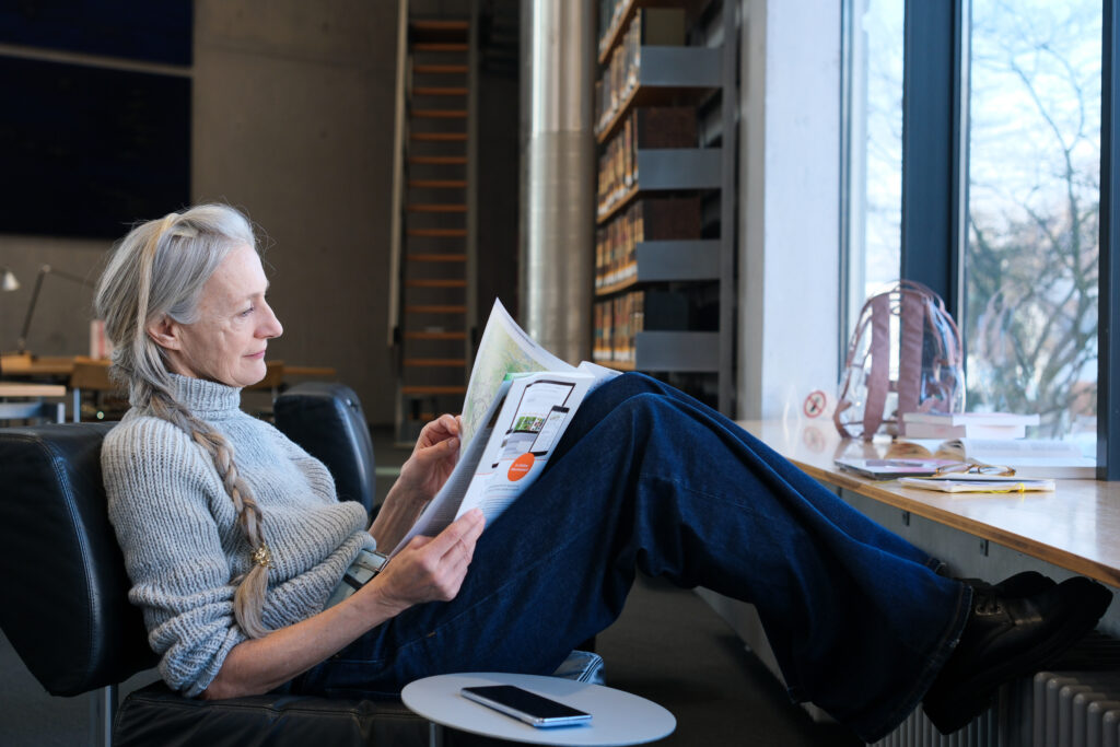 Eine Frau sitzt auf einem schwarzen Lederstuhl und liest konzentriert in einer Zeitschrift. Sie hat die Beine leicht angewinkelt und die Füße gegenüber auf einem Heizkörper abgestellt.